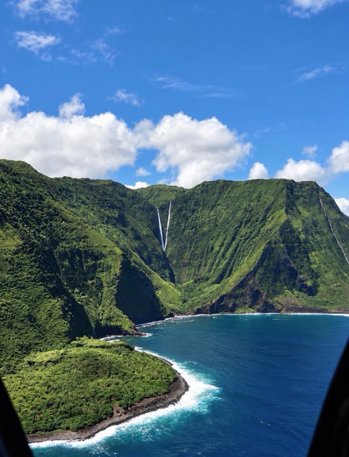 A blue cove below green cliffs and a thin waterfall