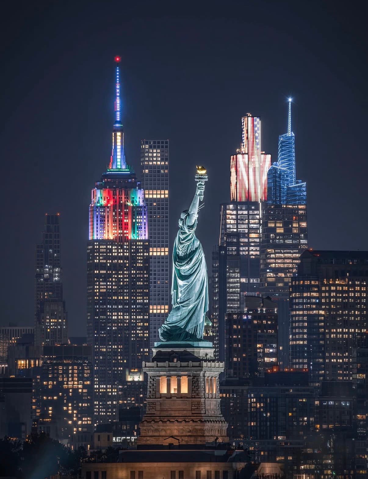 A night composite of the Statue of Liberty against Midtown's lit towers