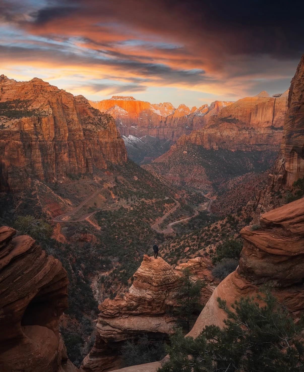 A hiker on a ledge above a deep red-rock canyon under a dramatic sky