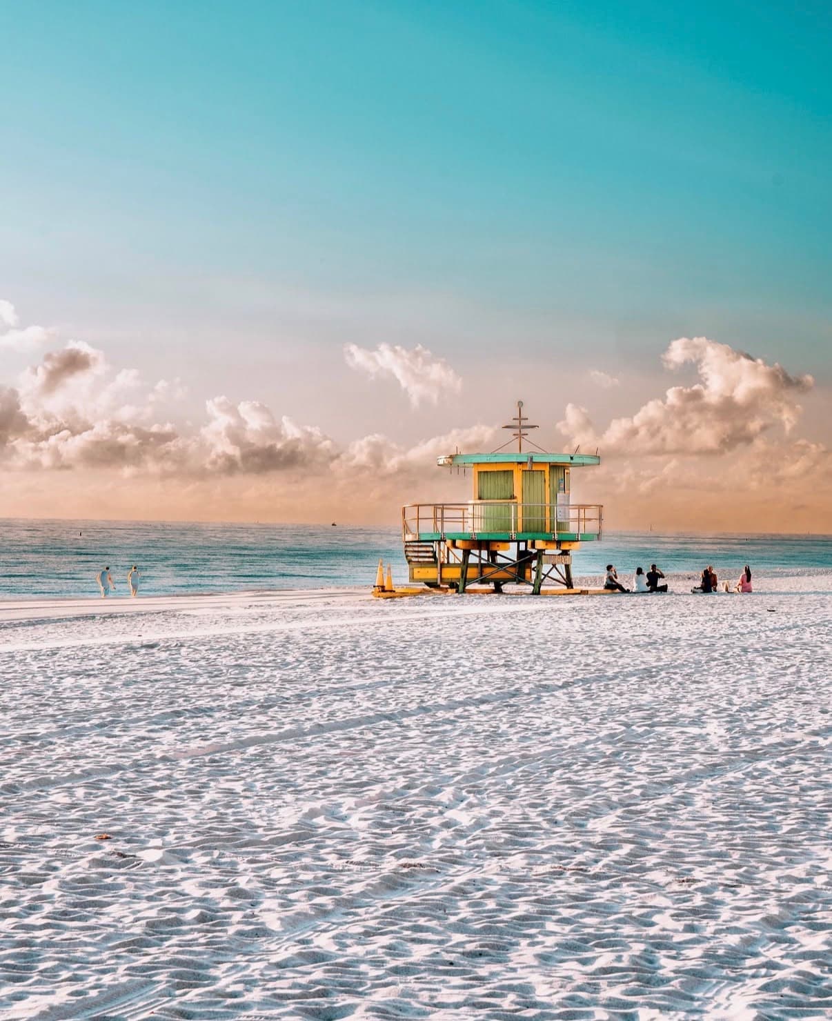 A pastel lifeguard tower on wide white sand below big clouds