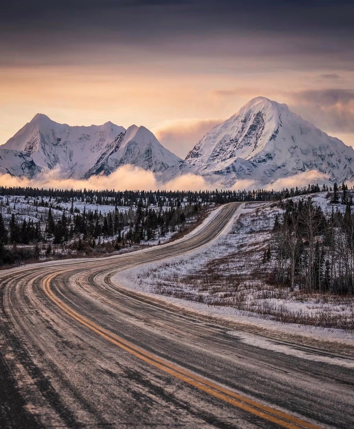 A winding highway through spruce and snow towards Alaska's peaks at dawn