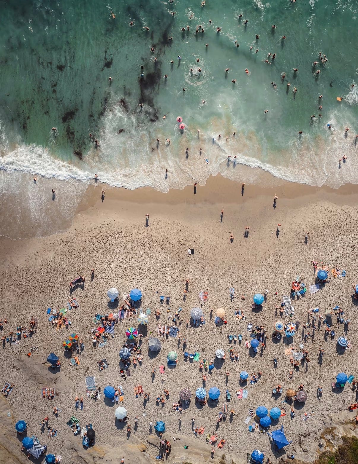 Swimmers and blue umbrellas on a beach seen from above, with surf at the water's edge