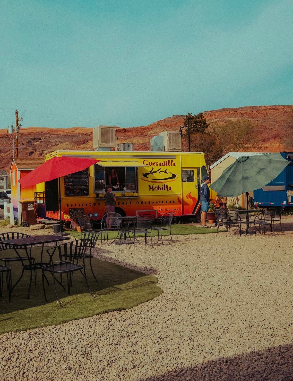 A yellow food truck with tables on a gravel lot under red-rock hills