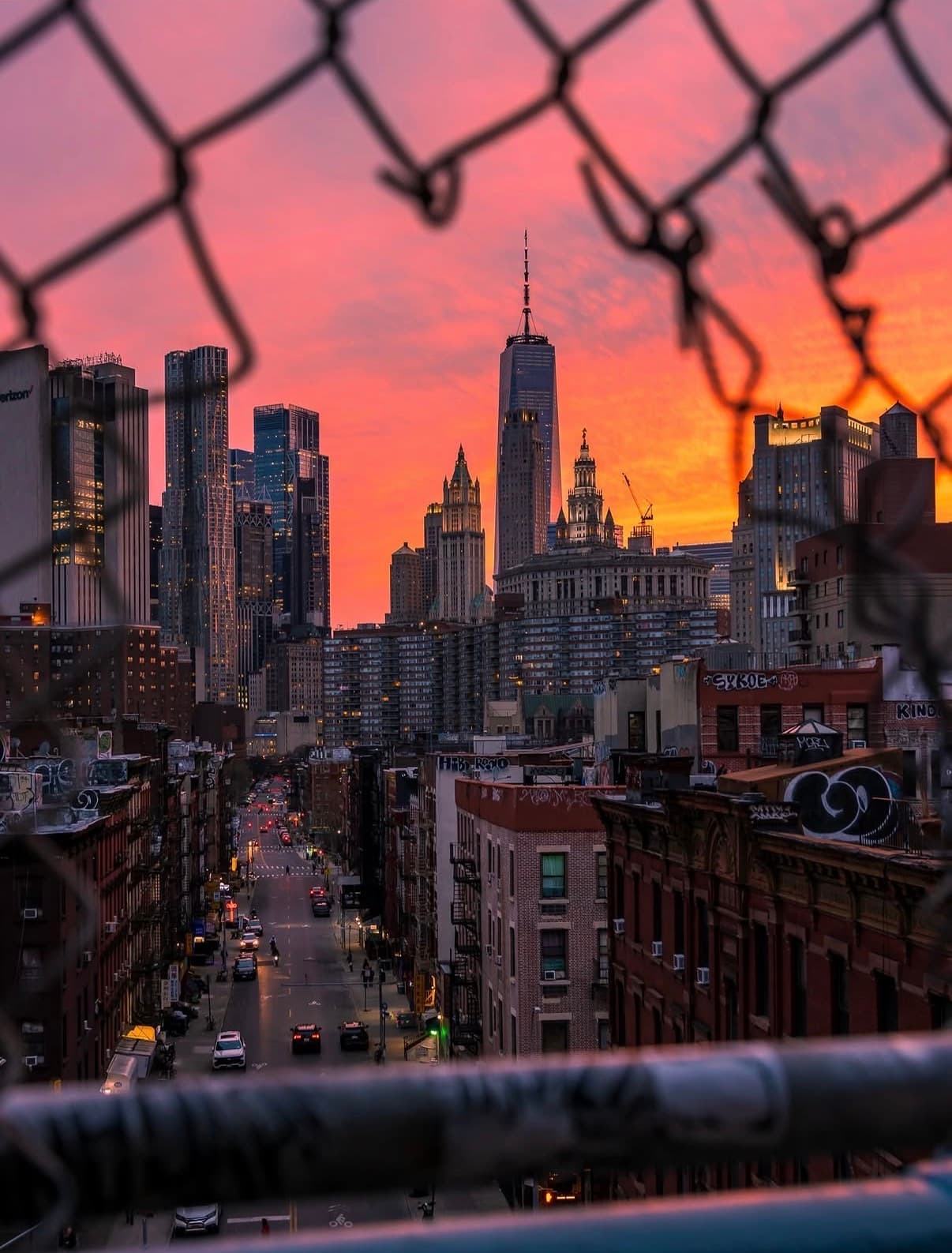 The Manhattan skyline at a pink sunset, seen through a chain-link fence